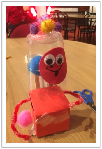 A red box body with a clear drink cup as a head. The face is a round red foam with googly eyes and sticking out tongue. It has two red and white striped pipe-cleaner arms. There are different coloured pom-poms on and in the head