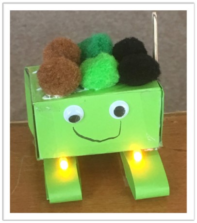 A green box with google eys, two rectangular box feet lit with yellow LEDs and pom-poms as hair