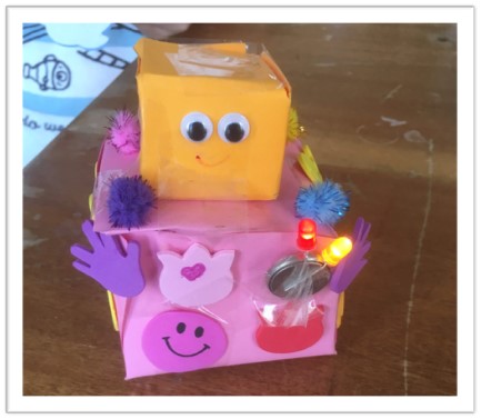 A pink box body and orange box head. The head has googly eyes and a smile. The body is decorated with foam shapes, LED lights and pom poms