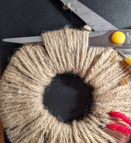 The scissors are positioned to between the wool and card on one side of the donut. It is in a position to start cutting the wool all the around the outer edge.