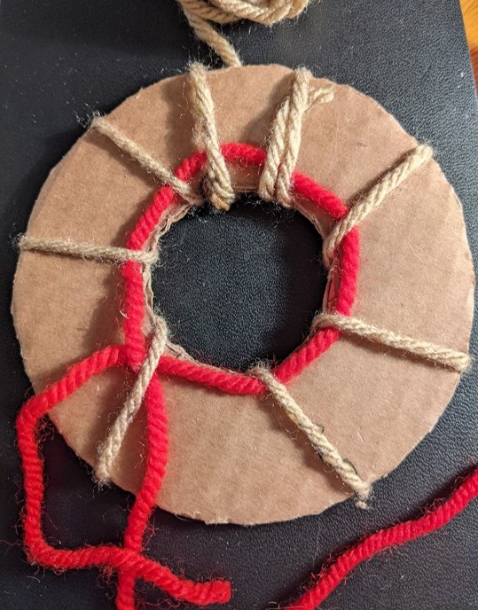 Brown wool has been looped around the donut at 8 different points to hold the tie thread in place. The tie thread follows the cardboard around the loop with a cross over before the ends are left hanging off the edge for tying together later